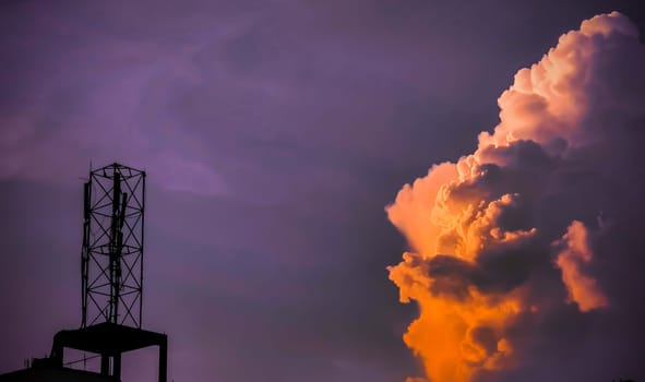 This image shows a cell tower silhouetted against a dramatic sunset sky. The sky is a vibrant purple and orange, with large, fluffy clouds. The tower is a tall, thin structure with a series of antennas at the top. The image is taken from a low angle, looking up at the tower. The contrast between the dark tower and the bright sky makes the image visually striking.