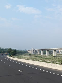 A view of a newly constructed elevated highway overpass, spanning over a lower-level road. The scene is set in a suburban area with a grassy field separating the road from the overpass. There are a few cars driving on the road, with a clear blue sky and light clouds in the background.