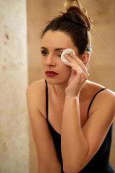 Beautiful young brunette woman washes away makeup in front of bathroom mirror