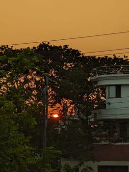 A sunset view from a residential neighborhood. The setting sun appears through the branches of trees, creating a warm glow in the sky. The image showcases a simple yet picturesque scene of everyday life in the city.