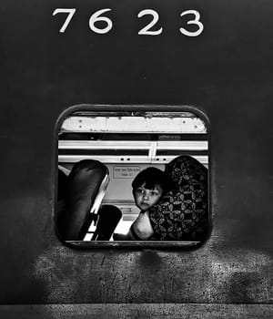 A young child peers out the window of a train car, their face illuminated by the light outside. The train car number, 7623, is visible above the window.