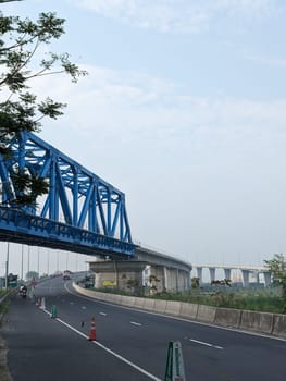 A view of a highway construction project with a blue steel bridge overpass. The road below is paved and empty, with traffic cones lining the edge. In the distance, another bridge can be seen. The sky is a light blue with some clouds.