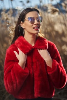 Beautiful young brunette woman in red fur coat poses in the meadow on sunny day