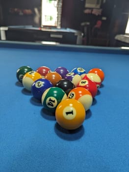 A close-up image of a triangle of pool balls arranged on a blue felt pool table. The balls are in a classic triangular configuration, with the cue ball at the front of the triangle, and the other balls numbered 1-15 in the background. The image captures the detail of the balls colors and numbers.