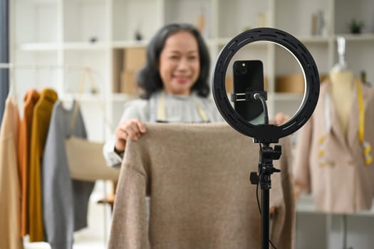 Smiling middle age woman showing clothes in front of smartphone camera while live streaming at her shop.