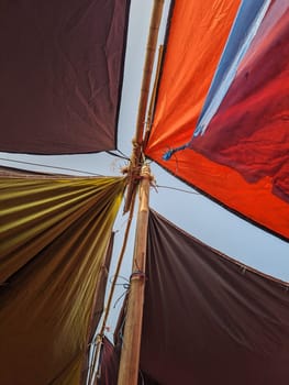A close-up shot taken from the inside of a shelter built using multiple brightly colored tarpaulins strung between bamboo poles. The canvas fabric creates a canopy overhead, allowing the viewer to see a glimpse of the sky.