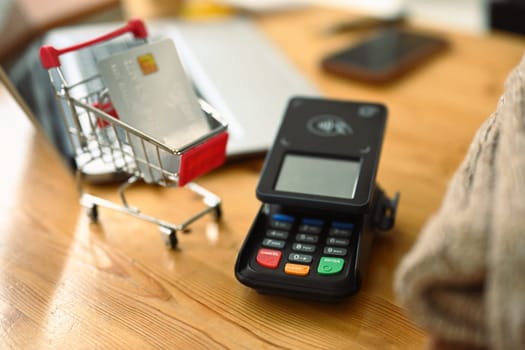 Modern bank payment terminal and shopping cart with credit card on wooden table.