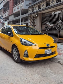A yellow Toyota Prius C is parked on a street in Dhaka, Bangladesh. The car is facing forward and the front of the car is in focus. The car is parked on a street with a building in the background.