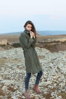 Confident woman in trench coat standing on rocky hillside with hands on hips overlooking scenic view