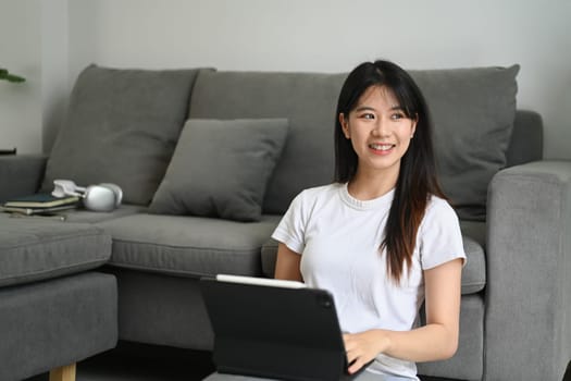 Charming young Asian woman sitting in living room with digital tablet and looking away.