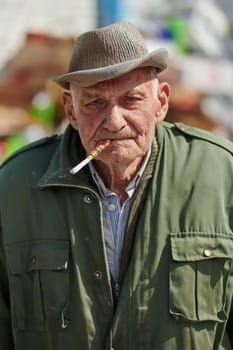 An elderly man is captured smoking a cigarette while wearing a dark green jacket and cap, exuding a sense of timeless style and sophistication.