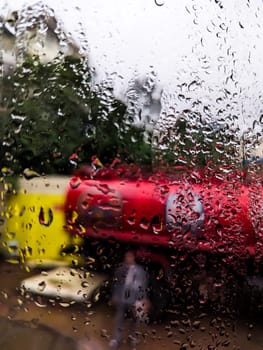 A blurry image of a red and yellow truck, likely a delivery vehicle, is seen through a window covered in rain droplets. The truck is parked in a city setting. The image captures the essence of a rainy day, with the raindrops blurring the background and creating a sense of movement. The image is taken from a perspective inside a vehicle or building.