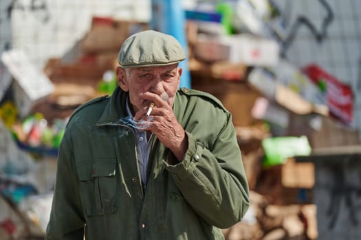 An elderly man is captured smoking a cigarette while wearing a dark green jacket and cap, exuding a sense of timeless style and sophistication.