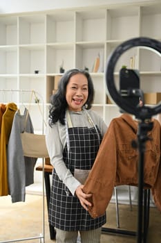 Cheerful senior lady small business owner selling clothes online by live streaming on mobile phone.