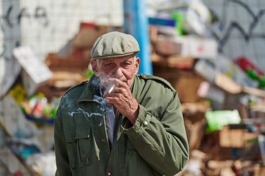 An elderly man is captured smoking a cigarette while wearing a dark green jacket and cap, exuding a sense of timeless style and sophistication.