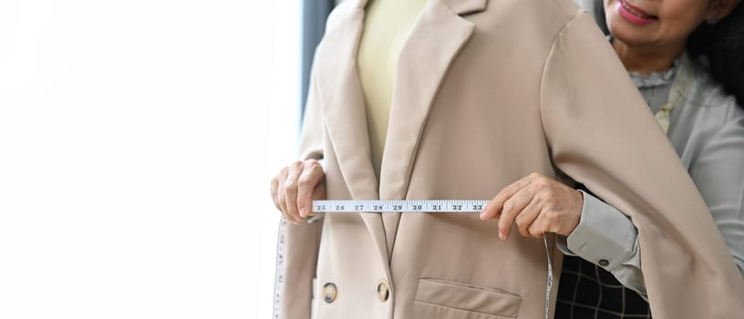 Skilled senior tailor working in the workshop studio measuring a jacket on a mannequin with tape measure.