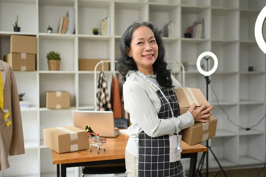 Confident senior woman small business owner holding parcel boxes of product and smiling to camera.