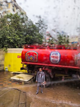 A man in a gray shirt and black pants stands next to a red tanker truck on a city street. The truck is parked in front of a yellow building. Rain is falling, and the scene is blurry due to raindrops on the camera lens.