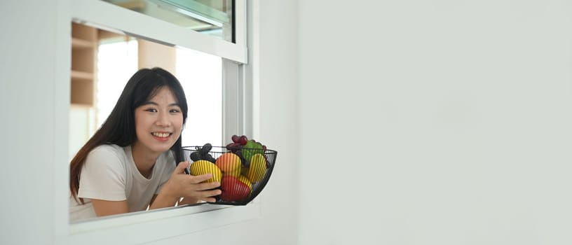 Cheerful young woman holding basket full of fresh fruit standing in kitchen snd smiling at camera, copy space for text.