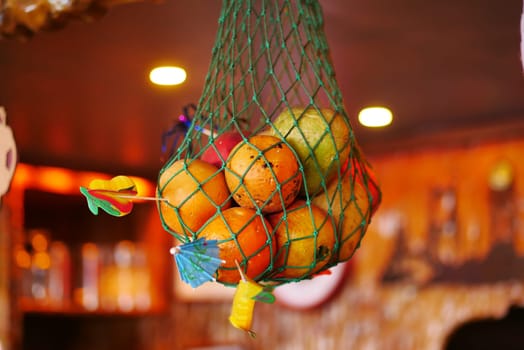 A vibrant, colorful assortment of fresh fruits hanging in a green mesh bag in a cozy interior setting.