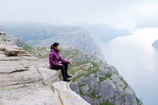 An Asian woman sits on the edge of the Preikestolen cliff in Norway, enjoying the breathtaking view of the fjord and the surrounding mountains.