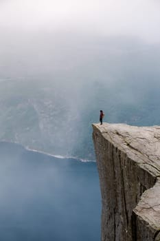 A lone hiker stands at the edge of Preikestolen, a dramatic cliff in Norway. The misty landscape and sheer drop below evoke a sense of awe and solitude.