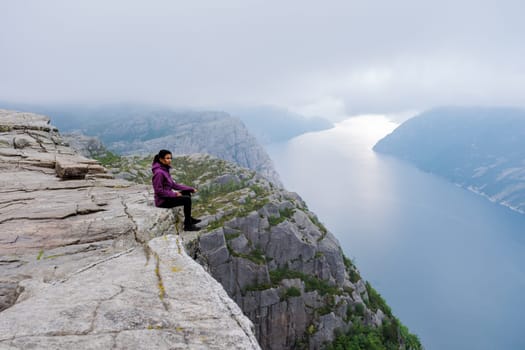 A Asian woman sits on the edge of the Preikestolen cliff in Norway, overlooking a stunning fjord. The misty morning adds to the dramatic landscape.