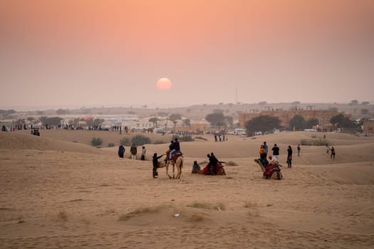 Jaisalmer, Rajasthan, India - 25th Dec 2023: line of camels carrying people tourists at sunset across thar desert in jaisalmer rajasthan showing a popular tourist activity