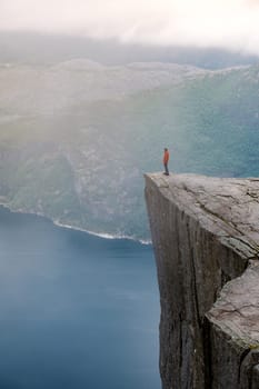 Preikestolen, Norway, A lone person stands on the edge of a sheer cliff overlooking a misty fjord in Norway. The vastness of the landscape is emphasized by the lone figure. Preikestolen, Norway
