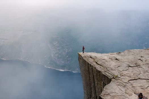 Preikestolen, Norway, A lone hiker stands on the precipice of a dramatic cliff in Norway, with a misty expanse of land and water stretching out before them.