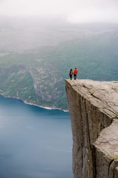 Two individuals stand on the edge of Preikestolen, a dramatic cliff in Norway, gazing out at the vast expanse of the fjord below. coupe of men and women at Preikestolen, Norway