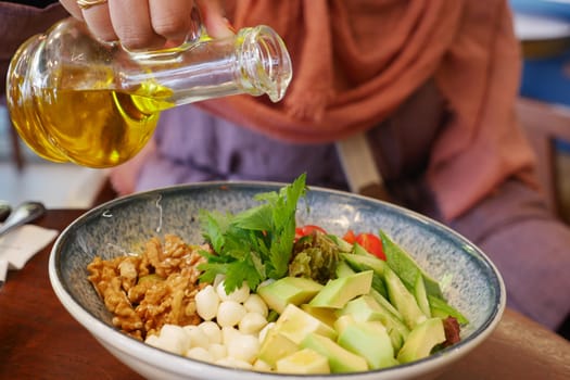 A person pouring olive oil onto a fresh, healthy salad, featuring avocados and walnuts, in a cozy restaurant setting.