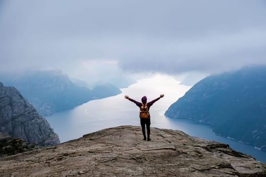 A lone hiker stands with arms outstretched on the edge of the famous Preikestolen cliff in Norway, overlooking a stunning fjord and the surrounding mountains. Preikestolen, Norway