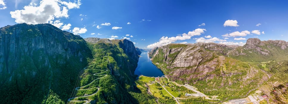 view of a winding road snaking through a stunning Norwegian fjord. Lush green mountains and a deep blue body of water create a breathtaking landscape under a clear blue sky. Lysefjord, Norway