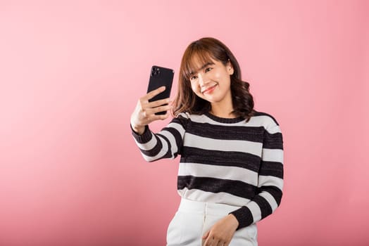 Happy Asian portrait beautiful cute young woman smiling excited making selfie photo, video call on smartphone studio shot isolated on pink background, female hold mobile phone raise hand say hello