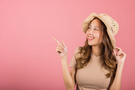 Portrait Asian young woman wearing straw hat is smiling and pointing finger up at an empty away side studio shot isolated on pink background, female people summer life enjoy in beach holiday concept