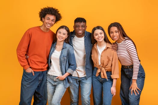 Group of five diverse young adults standing in front of a yellow background. They are all smiling and looking at the camera. The group appears to be friends and is enjoying each others company.