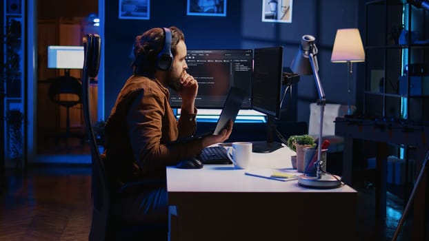 Engineer typing code in apartment office, using javascript programming language to develop software application. It admin using multimonitor desktop setup and notebook to finish tasks, camera B