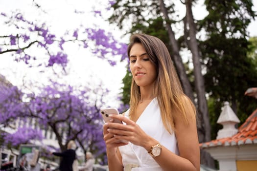 Happy young woman in white dress with brown hair using smartphone outdoors. Confident businesswoman text messaging via cellphone near violet Jacaranda trees in city park