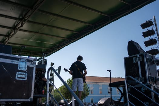 Back view of anonymous male artist playing the guitar on stage. Guitarist musician performs under blue sky in city concert setting.