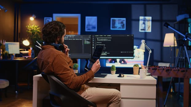 Software developer programming in home office, crosschecking lines of code between tablet and PC screen. IT admin holding portable device, using it as reference while coding on computer, camera B