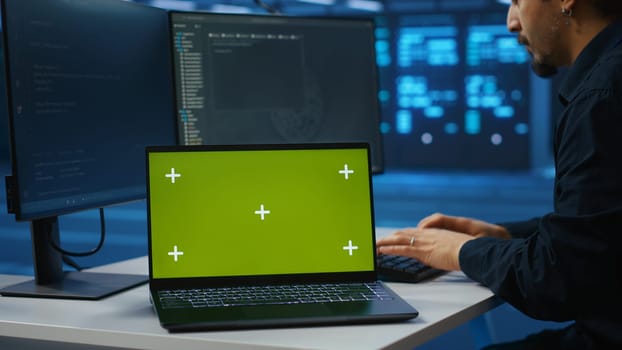 Isolated screen notebook in front of employee working in server farm used for processing data. Close up shot of mockup laptop on desk next to IT developer typing at PC, updating servers