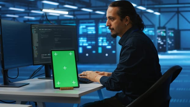 Man upgrading equipment in data center using isolated screen tablet. IT expert at PC desk in high tech workplace overseeing blade servers with chroma key device, panning shot