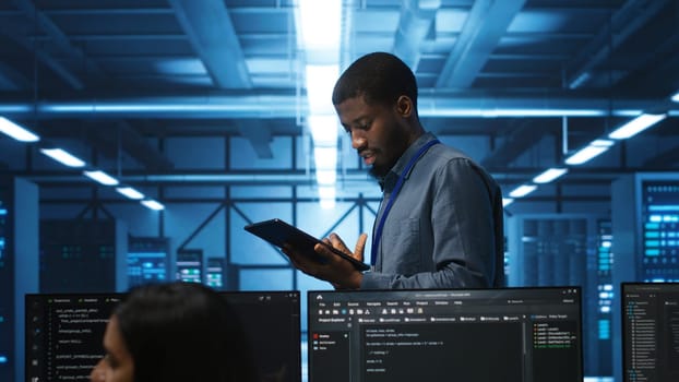 Engineer inputting data on tablet in server hub, monitoring data center energy consumption across hardware. Technician inspecting mainframes, ensuring malfunctions are repaired