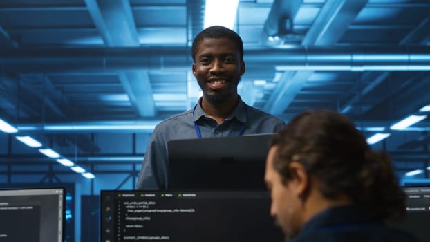 Portrait of cheerful african american manager supervising team in server room providing computing resources for workloads. Jolly supervisor oversees technicians in data center mending supercomputers