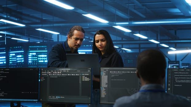 Multiethnic engineering team in server hub monitoring data center energy consumption across hardware using laptop. Multiracial colleagues inspecting supercomputers, ensuring malfunctions are fixed