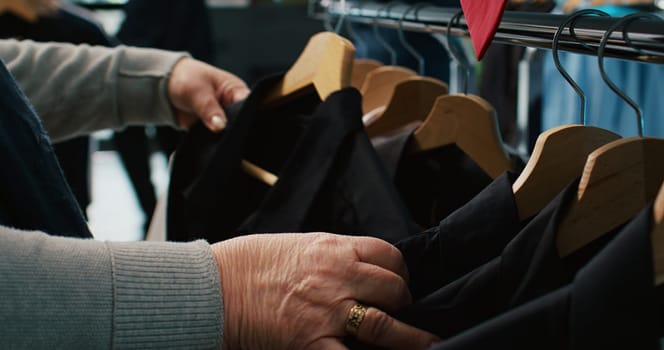 Senior woman browsing through hangers with elegant shirts, looking for new items to expand her wardrobe. Elderly customer in department store shopping for clothes. Close up. Camera B.