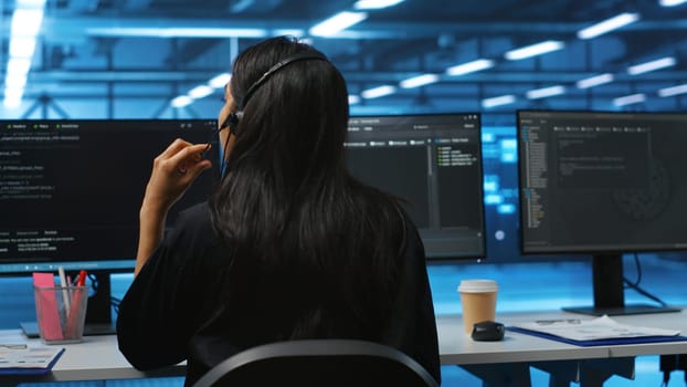 Computer scientist upgrading equipment in data center, wearing headphones to listen music. Programmer at PC desk in high tech establishment overseeing server rows, typing on keyboard