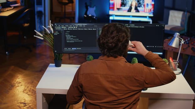 Computer scientist at home office desk writes binary code scripts and drinks from mug of coffee. IT expert in apartment enjoying tea while using programming to safeguard consumer data, camera B