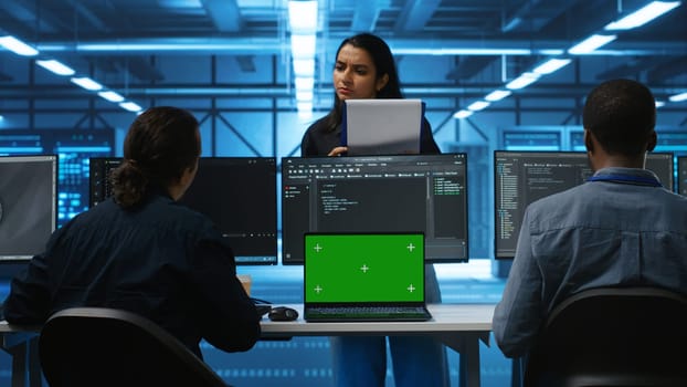 Multiethnic engineers working in high tech server hub, using green screen laptop and PCs to analyze data. Diverse employees examining infrastructure in data center, using chroma key notebook